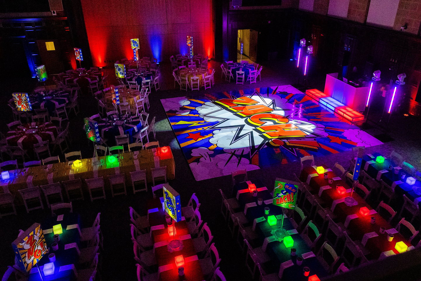 Comic-book-themed event room with a custom graphic dance floor, illuminated stage and coordinated centerpieces