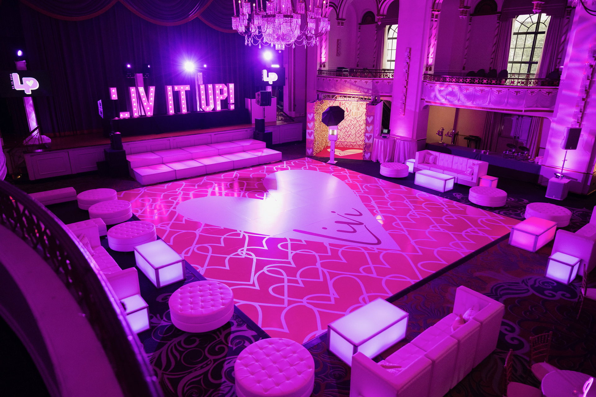 Pink heart-themed event ballroom with a custom dance floor, illuminated stage, lounge furniture and photo activation